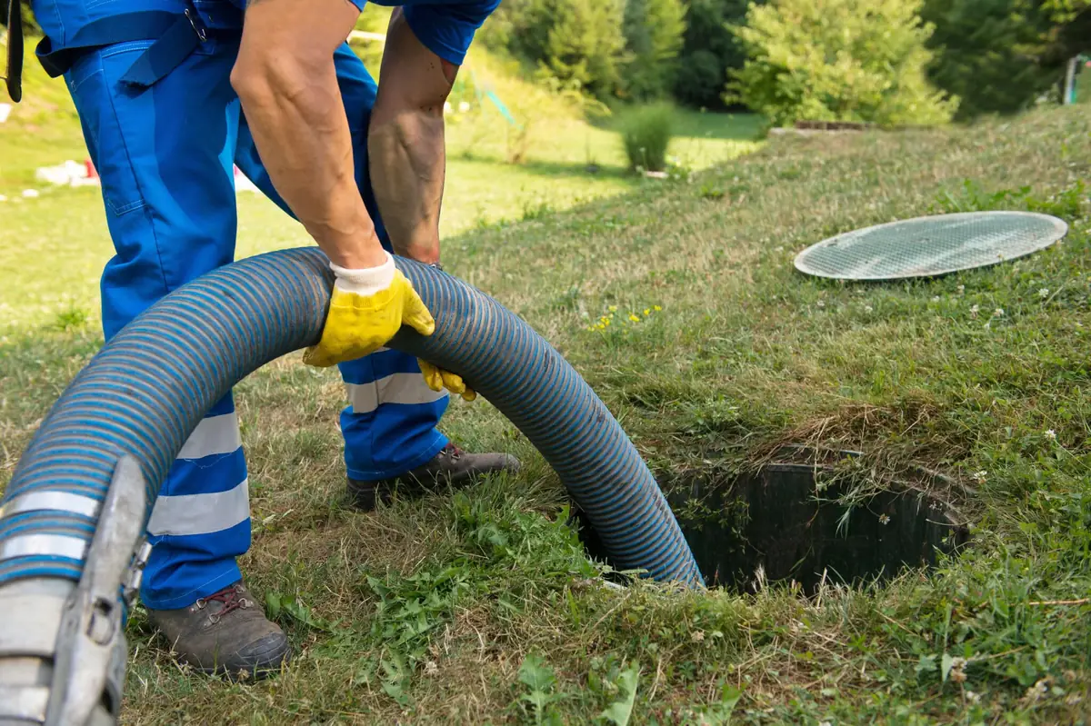 Cleanup worker in coveralls feeding a pump hose into an open sewer access in a yard